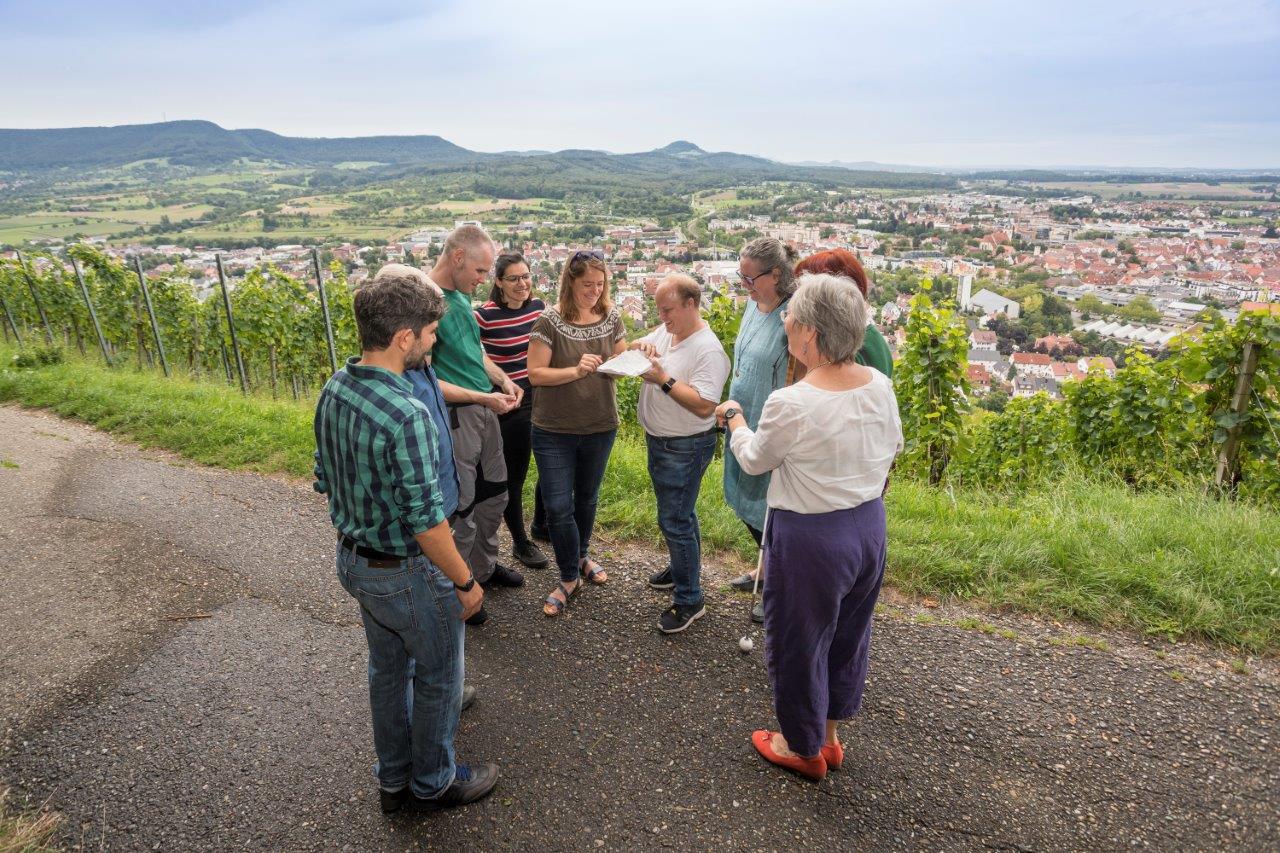 Eine tour de sens Gruppe steht an einem Aussichtspunkt oberhalb eines Weinbergs und eines Ortes und ertastet/betrachtet ein 3D Modell der Landschaft