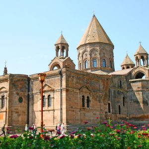 im Vordergrund blühen rote und pinke Blumen dahinter steht das Kloster Etchmiadzin vor einem blauen Himmel